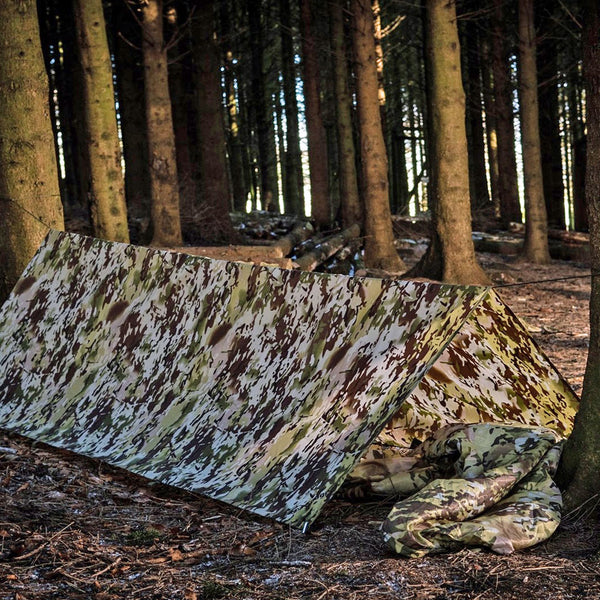 Lifestyle shot of a Snugpak Stasha G2 tarp in terrain colour forming a basic tent between two trees using paracord
