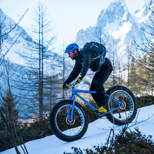 Lifestyle image of an adventure biker cycling on snow downhill in the Alps wearing a Sub Zero Factor 2 Plus mens long sleeve zip neck mid layer top underneath their lightweight down jacket